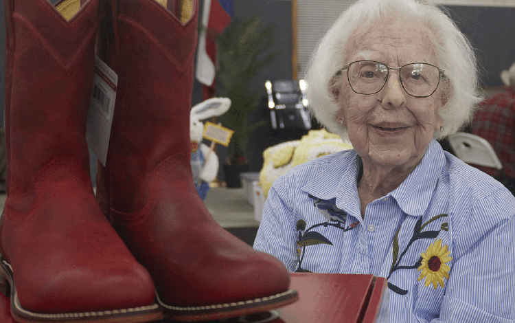 A lady with white hair, glasses, and a blue and white striped shirt with flowers posing and smiling next to her first pair of red cowboy roper boots. 